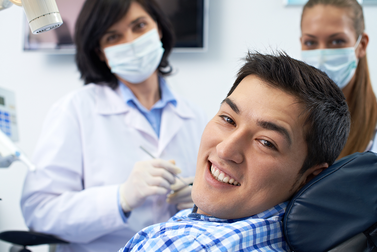 man-dental-patient-in-dental-chair-smiling-with-white-teeth-and-straight-smile-after-oral-care-with-smiling-dentist-and-assistant-in-background-at-dentist