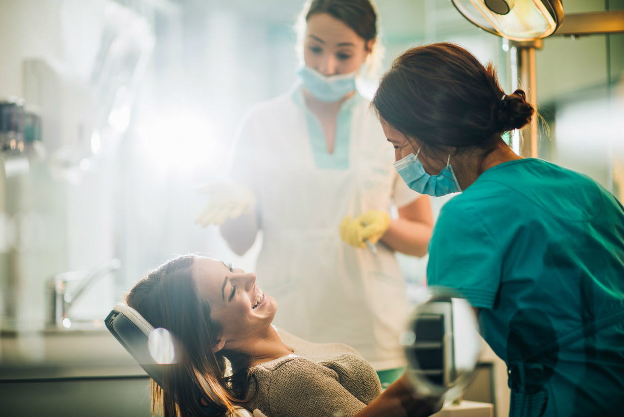 woman-dental-patient-in-dental-chair-smiling-while-discussing-sedation-treatment-with-smiling-female-dentist-and-assitant-at-dentist