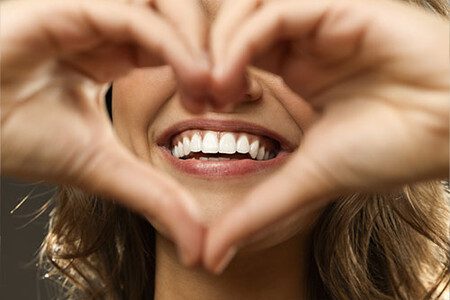 woman smiling with straight pearly white teeth peeking through her hands in heart shape